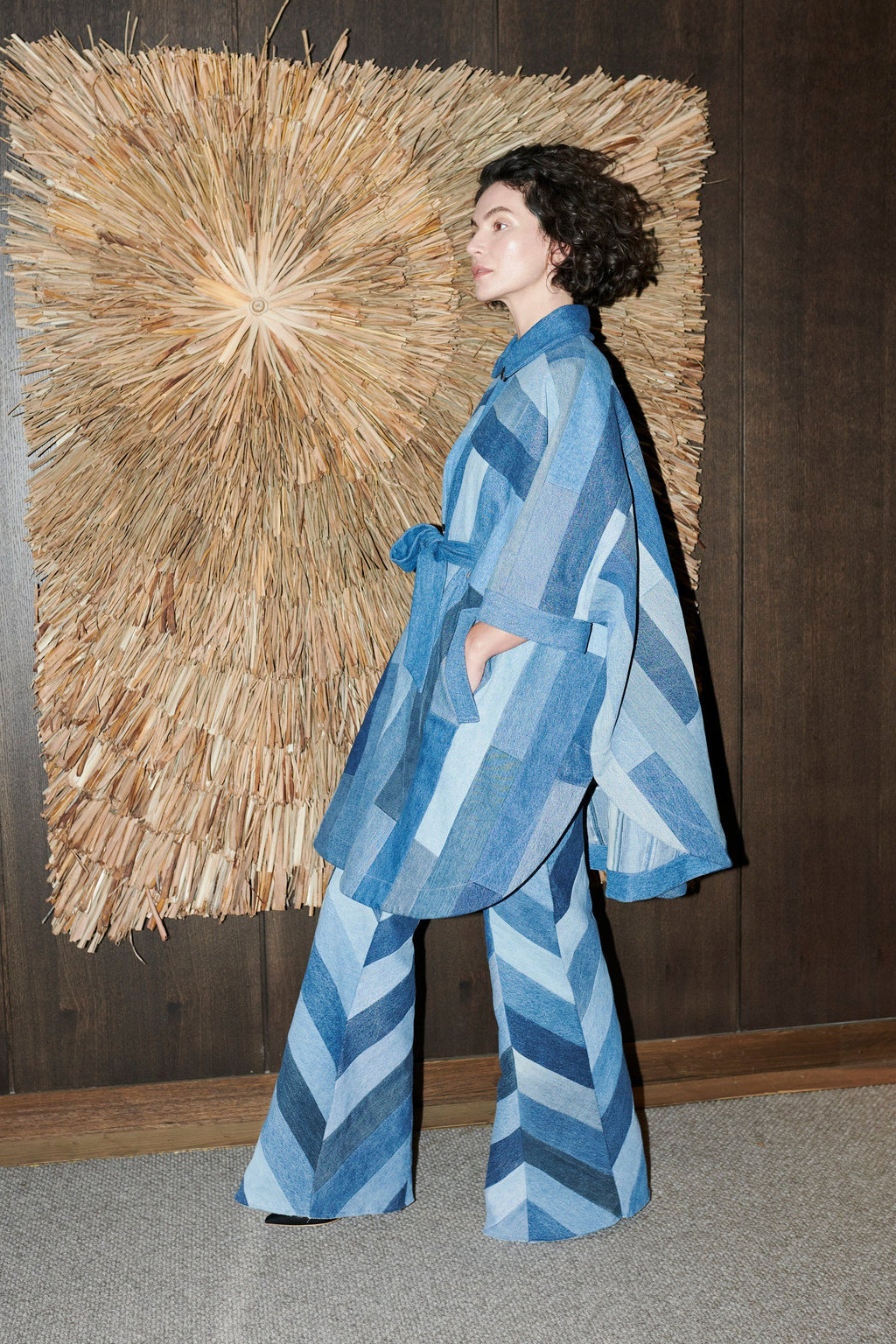 A model wearing a blue and gray patchwork denim two-piece set consisting of a long-sleeve belted jacket and matching wide-leg pants, standing in front of a dark wood-paneled wall with a large straw wall hanging.